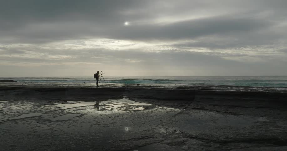 A silhouetted man films the breaking waves from a large tripod placed on a reflective wet rock platform under a dramatically cloudy sky