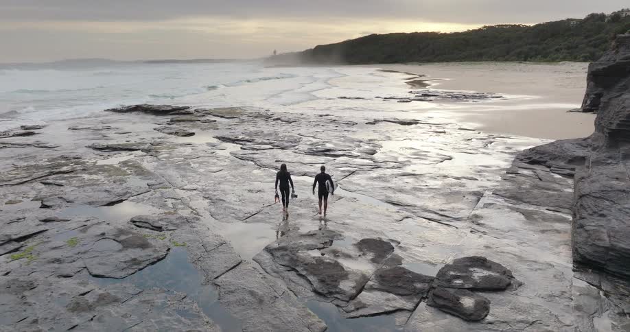 Two men in swimsuits walk along the rocky beach shoreline at midday one carrying a surfboard while waves can be seen in the distance hitting the shore