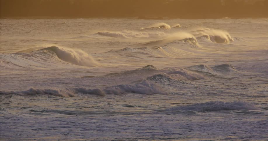 Powerful ocean waves break and spray in rows beneath dramatic golden sunset light