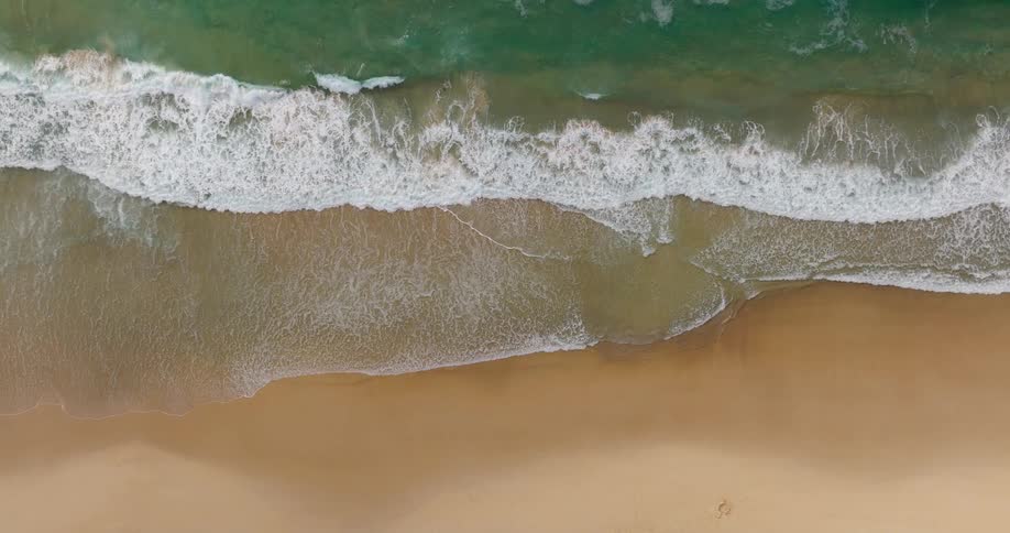 A direct overhead static shot captures the movement of multiple sets of clear turquoise waves crashing onto an expanse of smooth wet goldenbrown sand creating a thick layer of white foam that retreats and advances with the tide