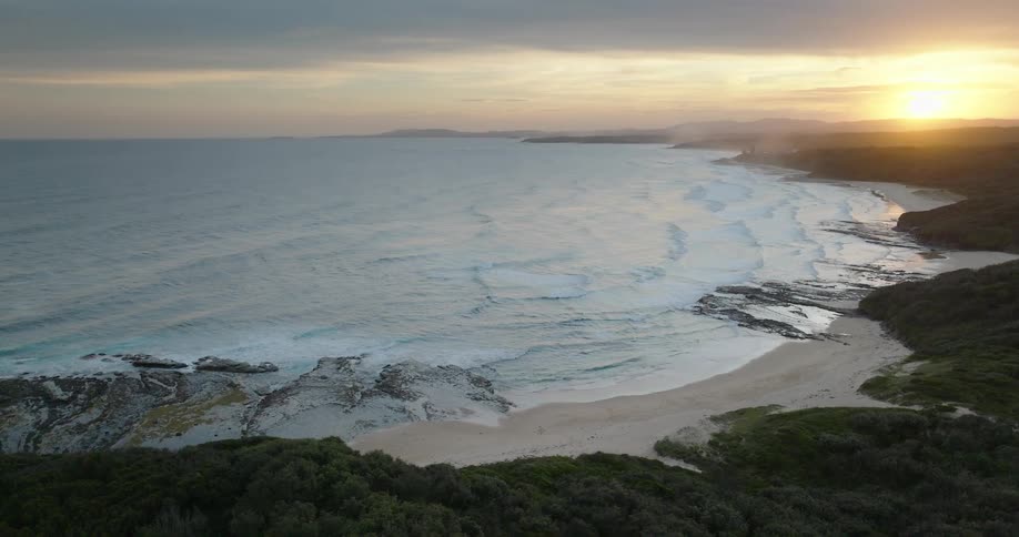 Waves roll in to shore and over the beach as the sun glows on the horizon