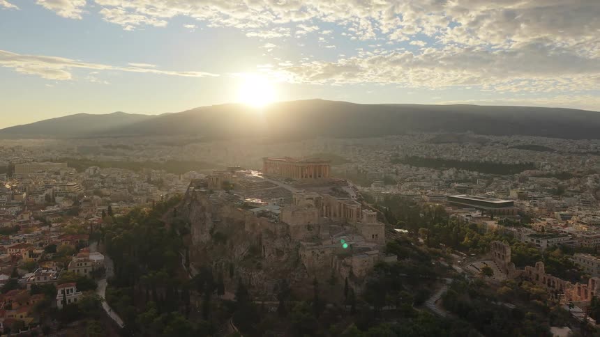 A low angle aerial view of the erechtheion temple on the acropolis showing the stone structure against a bright sunset sky and the city of athens in the valley below