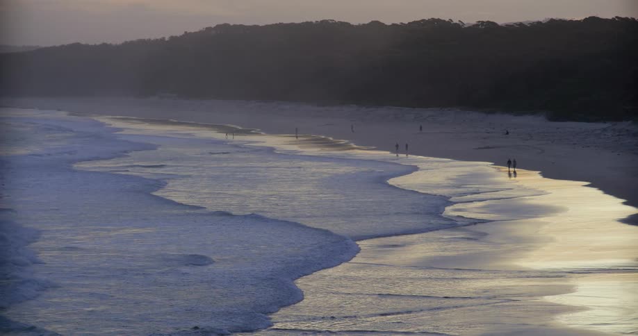 A few people are walking on a twilight beach featuring rolling waves meeting the sand with a dark distant forest forming the background