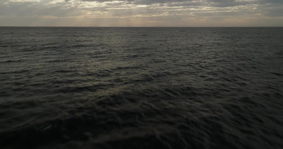 A camera flies steadily over dark choppy ocean waters moving toward a distant horizon where shafts of light break through a heavy layer of clouds