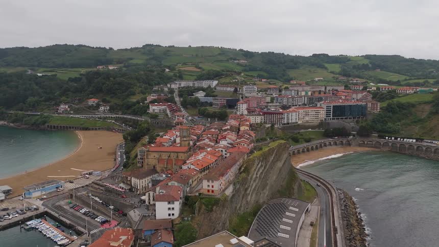 Cluster of redroofed buildings on elevated rock overlooks coastal town with marina and beach