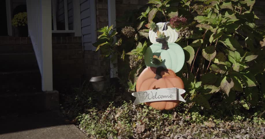 This is a static closeup shot of an autumnal decoration placed in front of a house next to a dark green shrubthe decoration consists of three stacked colorful pumpkinsorange at the bottom mint green in the middle and white at the topwith a bann