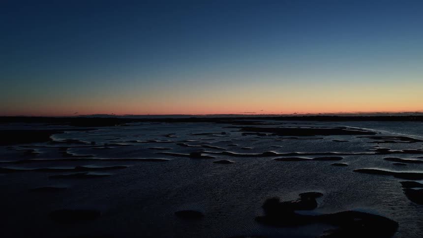 This is a static wide shot of an inlet or coastal marshland at twilightthe foreground is a dark reflective expanse of water and wet sand or mudflats contrasted against a calm gradient sky transitioning from deep blue to orange at the horizon