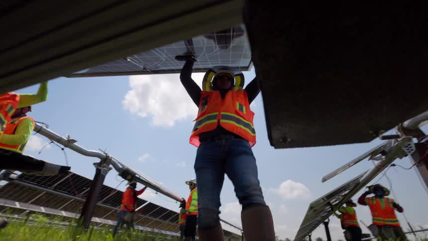 This is a lowangle shot of a group of workers in bright safety vests and hard hats installing solar panels on a sunny outdoor sitethe camera is positioned beneath one of the panels looking up as the workers lift carry and fasten the large solar r