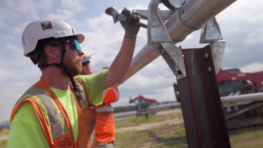 Two workers in vests and helmets install metal pipe on vertical support at construction site with vehicles in background