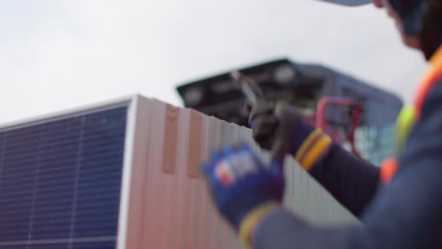 Person in vest and gloves handles solar panel at worksite with equipment in background