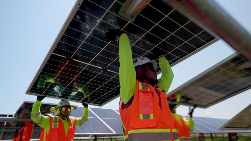 A team of construction workers in safety vests carry a large solar panel across a field and carefully install it onto a metal mounting frame