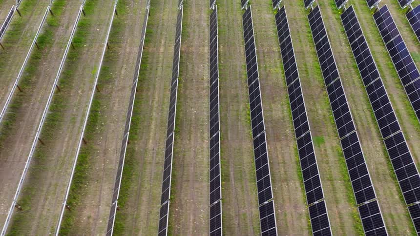 An aerial view moves slowly over a solar farm under construction showing long lines of installed panels next to empty metal support frames