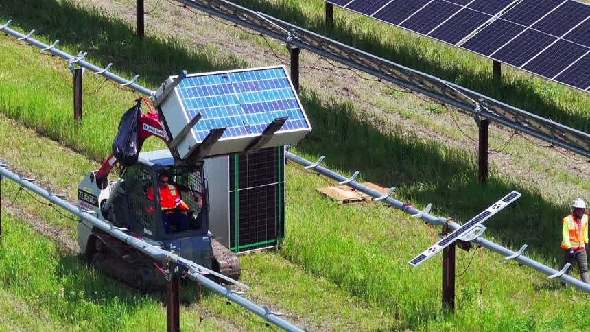 Solar panel lifted by hydraulic arm vehicle at solar farm operated by worker in orange vest