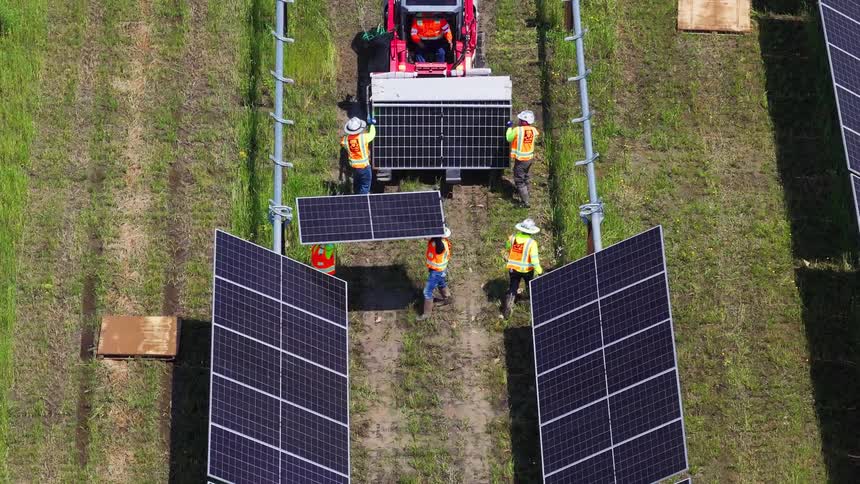 Red vehicle with solar panel is surrounded by six workers in grassy field with structures being installed
