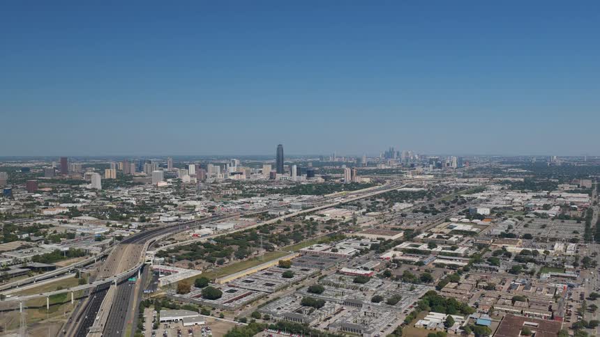 A wide aerial view shows a sprawling cityscape with highways leading toward a distant skyline under a clear blue sky