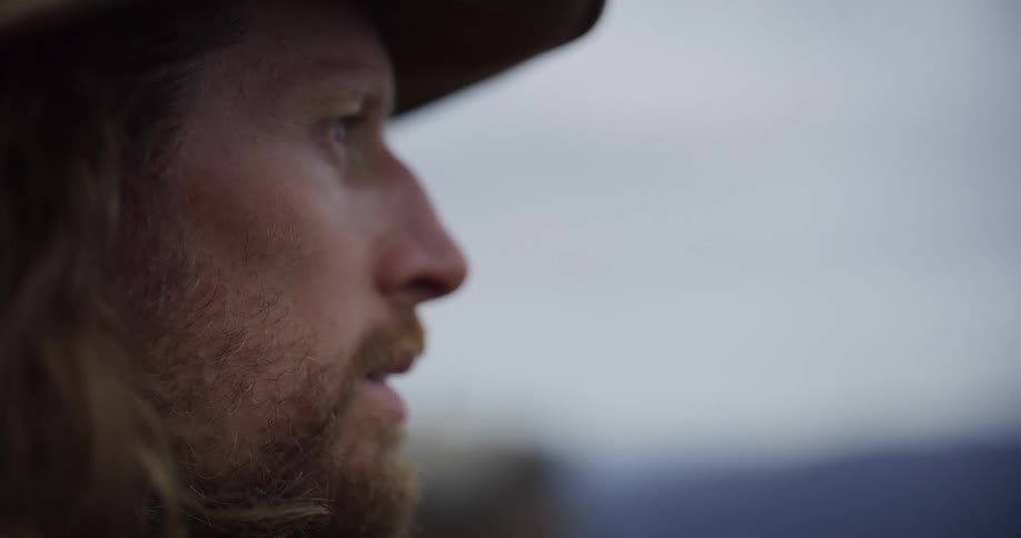A closeup shot of a mans face in profile showing his reddishblond hair beard and a partial view of a hat he is wearing he is looking off into the distance with a serious or pensive expression against a blurred overcast background