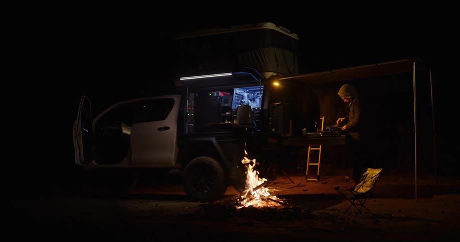 Night campsite with an equipped pickup truck fire burning on the ground and a person cooking beside the vehicle