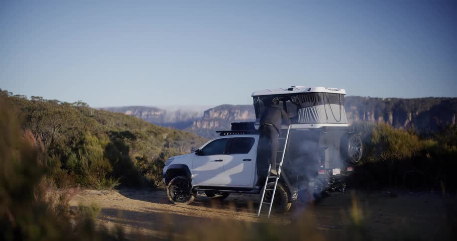 Man climbs down a ladder from the tent on the top of a vehicle at a campsite in the wilderness during the daytime