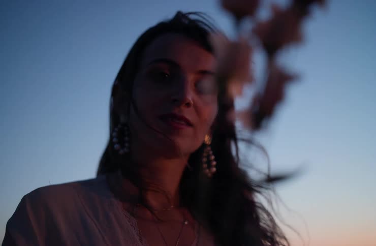 The face of a woman holding two flowers with pink petals in her hand set outdoors with a blue sky in the background