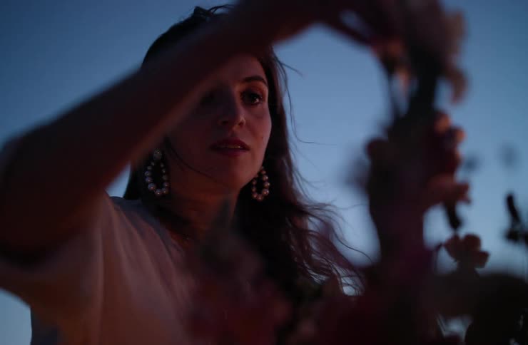 A closeup lowangle shot shows a woman arranging light pink flowers against a deep blue twilight sky she is silhouetted and her face is dimly lit with her long dark hair blowing slightly in the wind