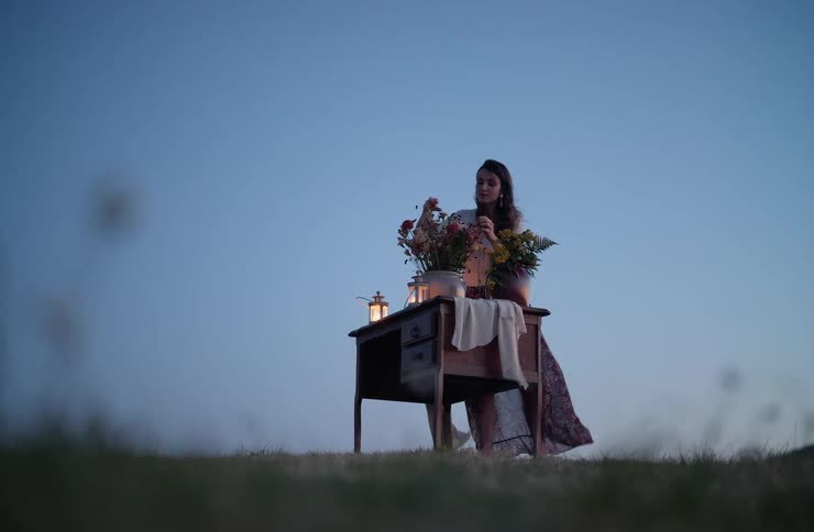 A woman in a long dress arranges flowers on a wooden desk with a lit lantern in a grassy field at dusk