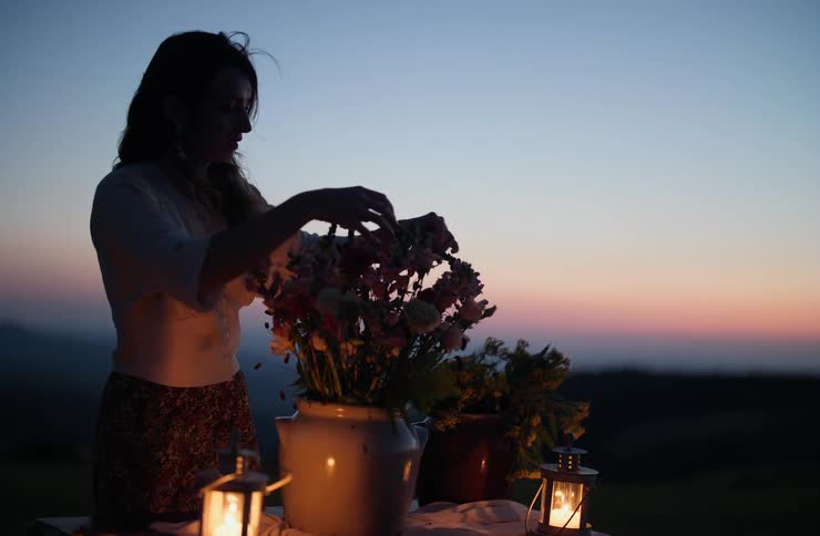 A woman arranges flowers on a table lit by lanterns against a twilight sky