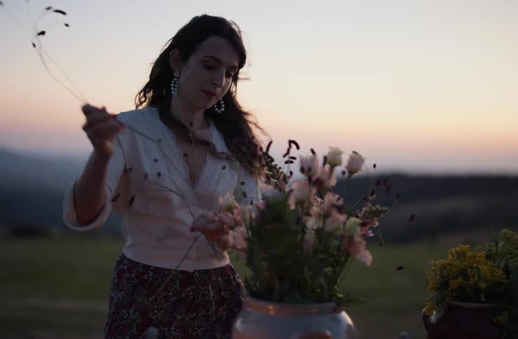 A woman is seen arranging flowers in a large vase outdoors during a sunset or twilight setting she is carefully placing stems of small dark flowers into an arrangement that already contains light pink and green flowers