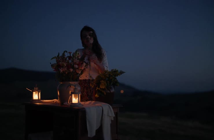 A woman is arranging a bouquet of flowers on a table with two lit candles in an outdoor setting at night