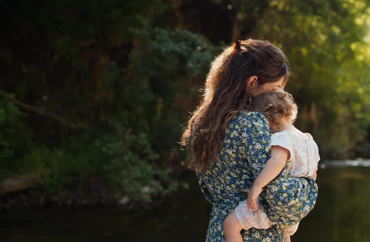Woman in blue floral dress holds child with pink accents beside stream surrounded by greenery