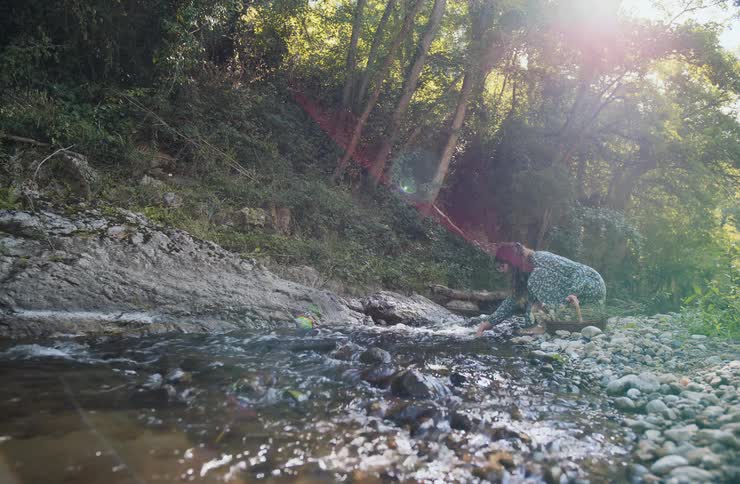 A woman wearing a patterned dress walks to the edge of a rocky stream in a lush sunlit forest she kneels down on the pebblecovered bank and reaches her hand into the clear running water