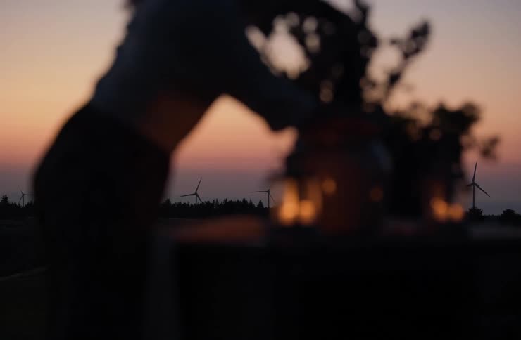 Blurred silhouette of a woman leaning over a table with flowers and candles against a sunset horizon with wind turbines