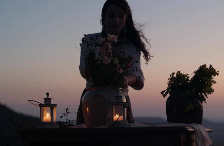 A woman is arranging flowers in a vase outdoors at dusk there are two lit lanterns and a second vase of yellow flowers on a table in front of her set against a sunset or twilight background over hills