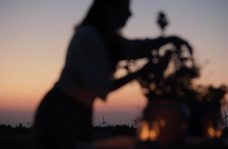 A woman is silhouetted against a colorful sunset sky arranging flowers in a vase on a table in the background wind turbines are visible on the horizon suggesting an open possibly rural settingthe scene is soft and blurred with light from what a