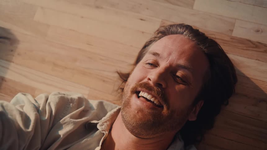 A young man with facial hair and long hair lies on a wooden floor indoors laughing and having fun