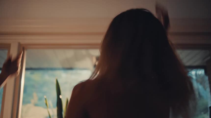 A young woman with long blonde hair is shown dancing exuberantly tossing her head and moving her arms in a sunlit roomshe is wearing a light top and appears to be enjoying the moment with another person whose arm is visible next to her