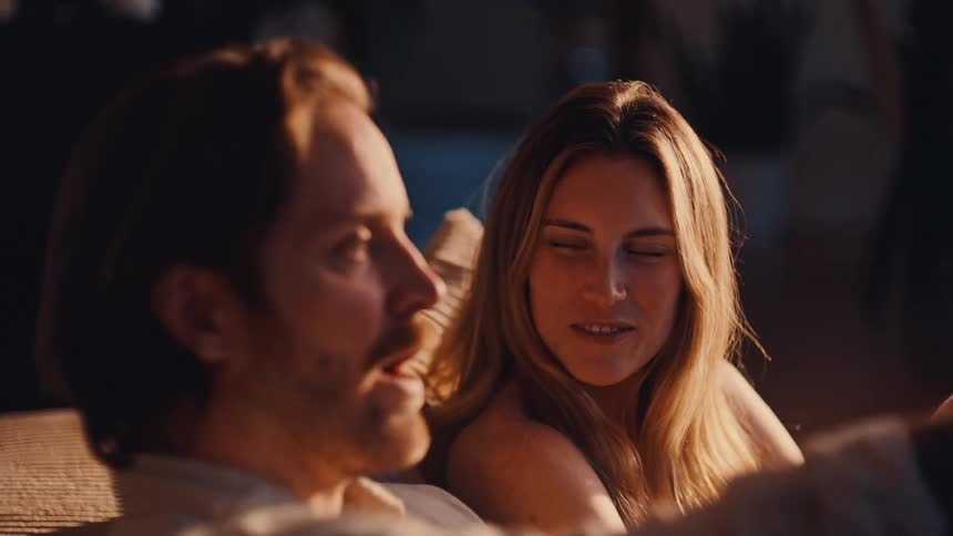 Two people sit together in cozy indoor setting sharing an intimate moment under soft lighting
