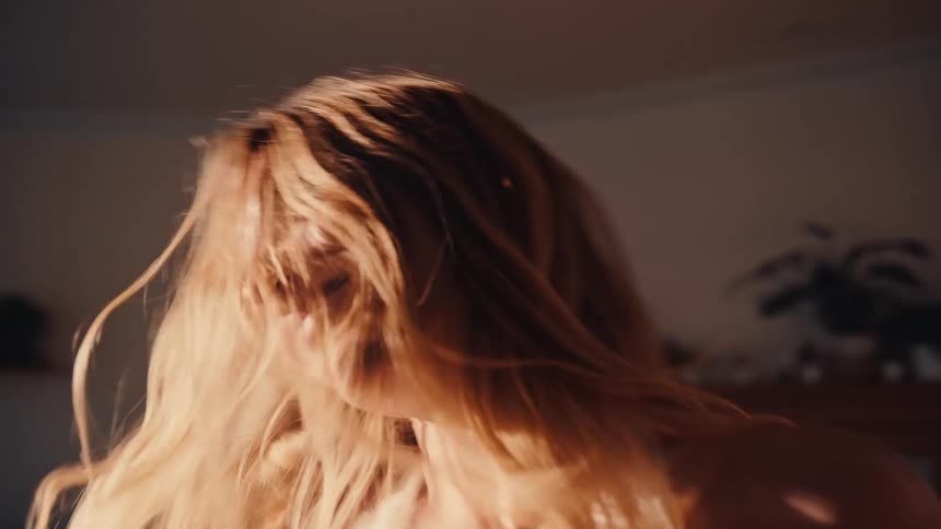 A young woman with long blonde hair dances in her living room sunlight streaming through the window onto her face as she enjoys herself