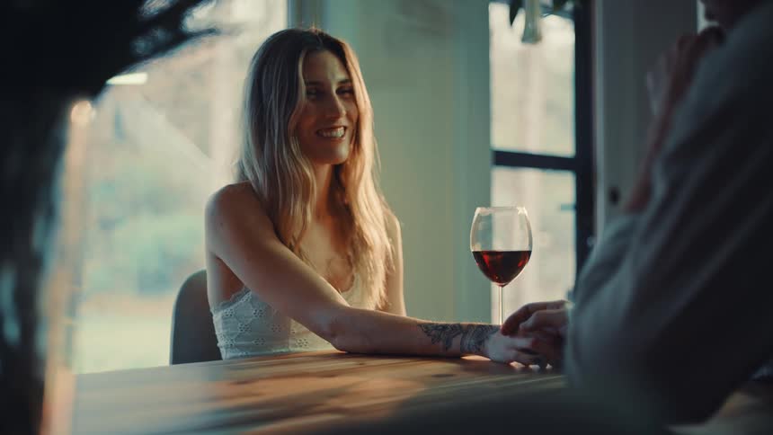 A smiling woman holds hands with a man across a wooden table next to a glass of red wine