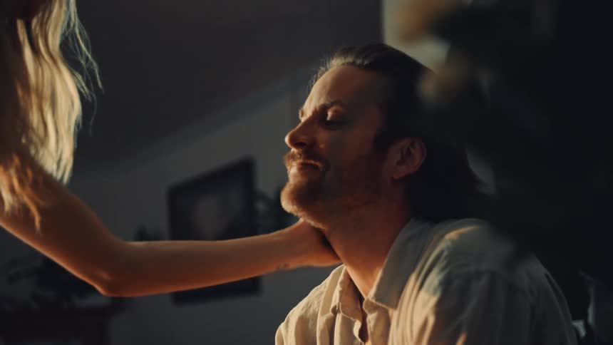 A young couple kiss indoors as sunlight reflects into the room with the man sitting and the woman standing beside him