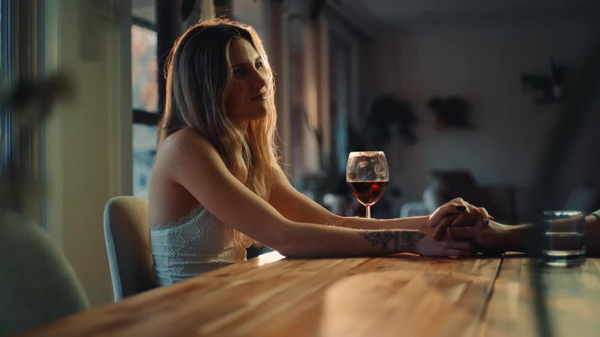 A young woman sits at a dining table with a glass of wine in front of her placing her hand on the table as someone holds it down while she stares at them