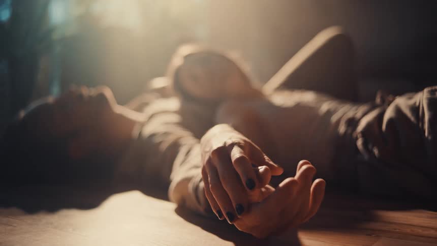 A young couple lie on the floor indoors with the woman on top of the man as they hold hands sunlight streaming through the window into the room