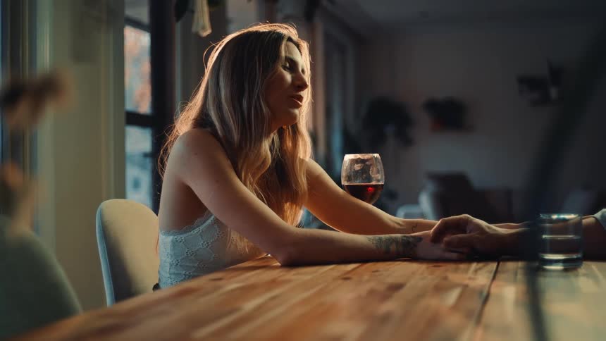A blonde woman wearing a lightcolored lacy top is seated at a wooden table across from another person out of frame bathed in warm intense light coming from behind hershe is holding the other persons hand on the table maintaining eye contact an