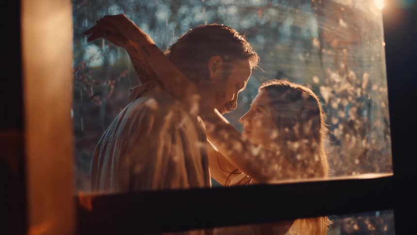 A couple stand outdoors beside a glass window hugging and kissing as rain drips from the roof with sunlight streaming down from the sky