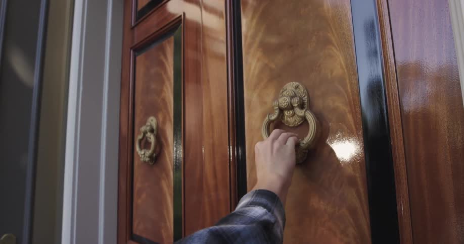 Close up of a person knocking on a large wooden door at a building entrance during daytime