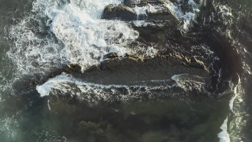 The first video shows an extreme overhead view of rough dark rock formations being continuously hit by white crashing ocean waves and seafoamthe second video shows two small figures walking across a vast uniform textured gray landscape viewed from