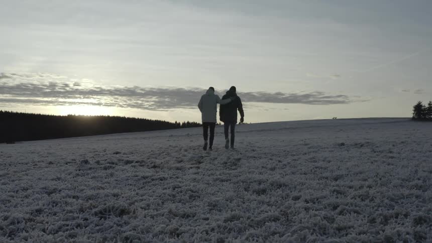 Two men walk together through a vast frosty field toward the rising sun and one gently places his arm around the others shoulder