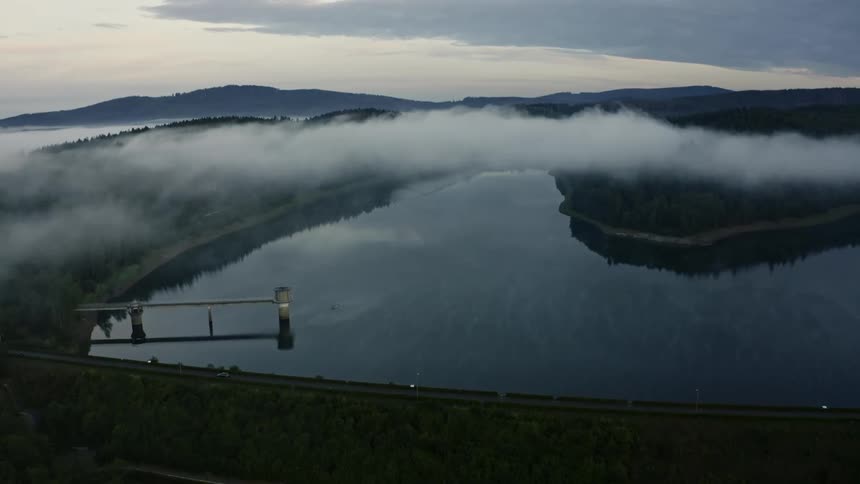 Clouds hanging over forested mountains along the coast with a small boat on the water and a car on the road