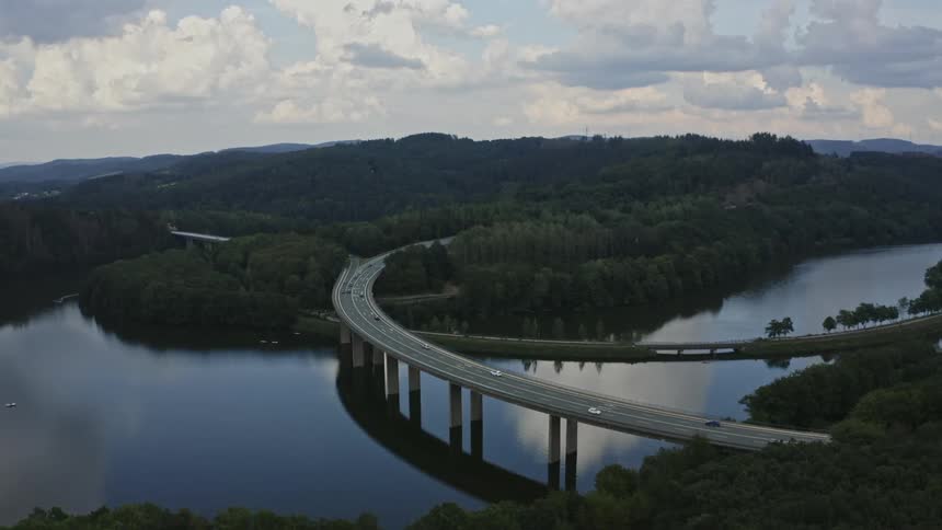 An elevated wide shot frames a concrete highway bridge with traffic curving over a dark still body of water flanked by densely forested hills on either side with clear reflections of the structure and the cloudy sky