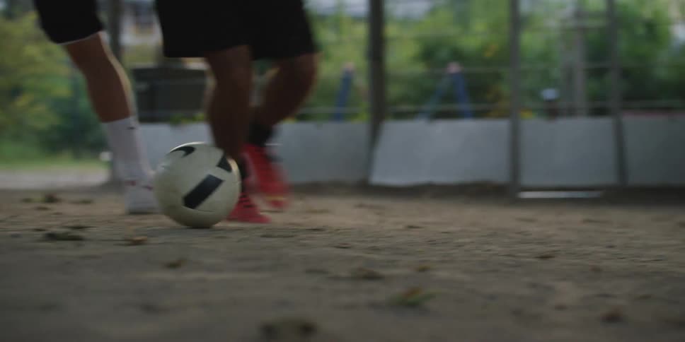 The video shows a closeup of two peoples legs and feet primarily focusing on one person wearing bright orange and black football boots controlling a soccer ball on a dirt groundthe other person wearing white socks and white sneakers appears to be