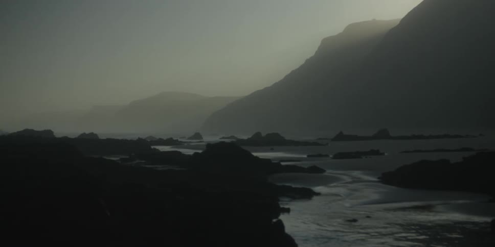 A misty coastal landscape reveals dark rocky shores and towering mountains at dawn